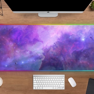 May include: A rectangular, large mouse pad with a vibrant nebula design in shades of purple, pink, and blue. The pad has a glowing, multi-colored LED edge. A white keyboard, mouse, and coffee cup are on a wooden desk.