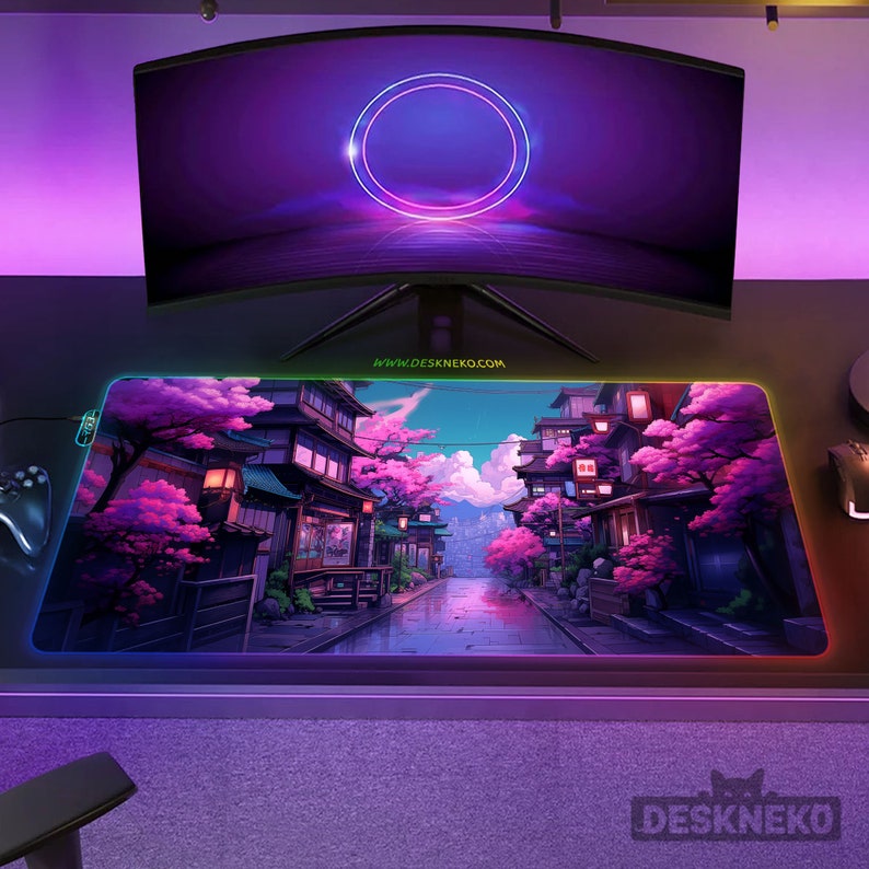 May include: A rectangular gaming mouse mat featuring a Japanese landscape with pink cherry blossom trees and traditional buildings. The mat has a colourful LED edge and the text "DESKNEKO.COM". A curved monitor is above the mat.