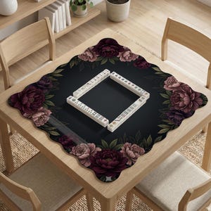 May include: A wooden table with a black mat featuring a floral design and a set of Mahjong tiles. The mat has a dark background with burgundy and pink peony flowers and green leaves. The Mahjong tiles are arranged in a square.
