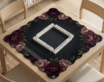May include: A square wooden table with a black mat featuring a floral design and a set of Mahjong tiles. The mat has a dark background with burgundy and pink peony flowers and green leaves. The Mahjong tiles are arranged in a square.