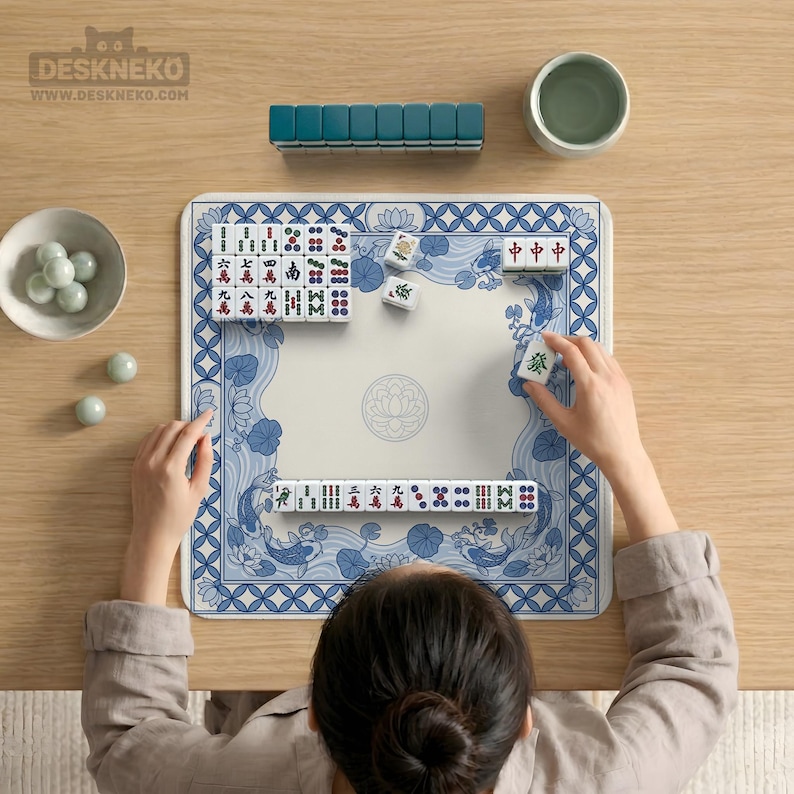May include: A person playing Mahjong on a blue and white patterned mat. The game set includes tiles, a rack, and a small bowl. The mat features a floral and koi fish design. The game is set on a wooden table.