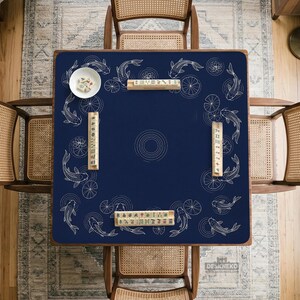 Puede incluir: Un mantel de mesa de Mahjong azul marino con un dise&ntilde;o de carpas koi y nen&uacute;fares. El mantel cuadrado tiene un patr&oacute;n circular central. Hay fichas de Mahjong y un cuenco blanco peque&ntilde;o sobre la mesa. Cuatro sillas de madera rodean la mesa.