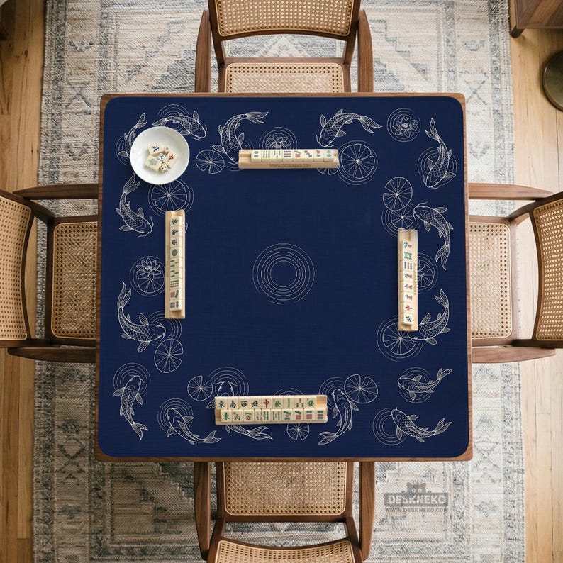 Puede incluir: Un mantel de mesa de Mahjong azul marino con un dise&ntilde;o de carpas koi y nen&uacute;fares. El mantel cuadrado tiene un patr&oacute;n circular central. Hay fichas de Mahjong y un cuenco blanco peque&ntilde;o sobre la mesa. Cuatro sillas de madera rodean la mesa.