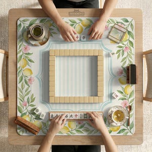 May include: A mahjong game in progress on a wooden table. The game board features a floral and lemon design with a light blue striped border. Players are arranging tiles, with cups of tea and accessories nearby.