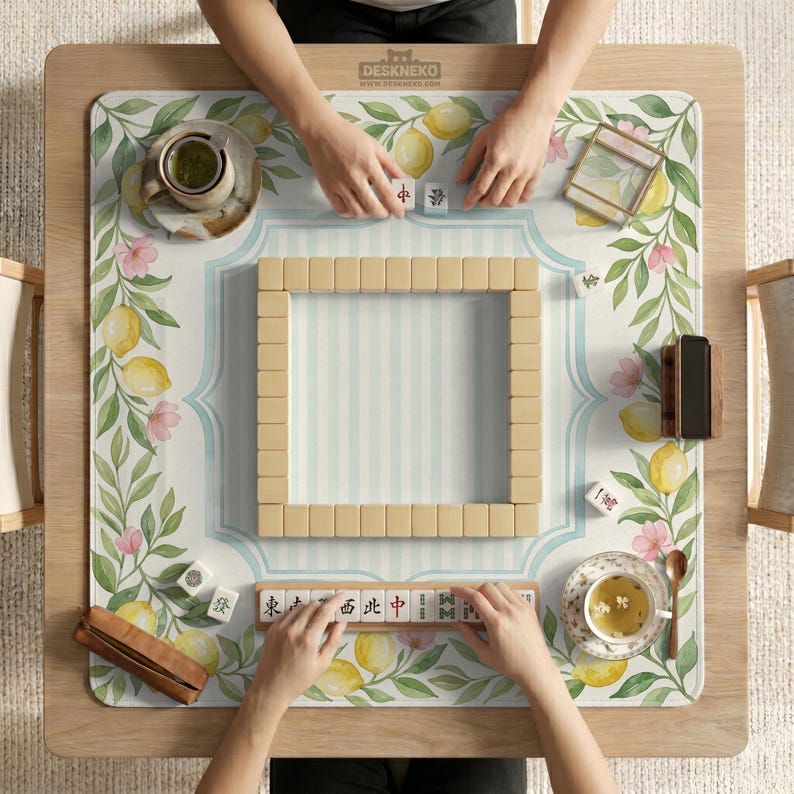 May include: A mahjong game in progress on a wooden table. The game board features a floral and lemon design with a light blue striped border. Players are arranging tiles, with cups of tea and accessories nearby.