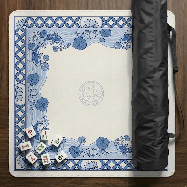 May include: A white Mahjong mat with a blue and white koi fish and lotus flower design. The mat has a decorative border and a central lotus flower emblem. A black carrying case is on the right side of the mat. Mahjong tiles are visible.