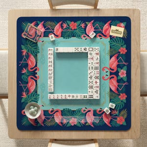 May include: A square mahjong game board with a teal center and a navy blue border. The border features pink flamingos, tropical leaves, and red flowers. White mahjong tiles are arranged around the teal center. The board is on a light wood table.