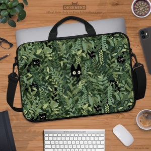 Peut inclure: Sacoche pour ordinateur portable avec une poignée noire et une bandoulière. Le sac présente un motif de feuilles vertes avec des visages de chats noirs qui regardent. Le sac est sur un bureau en bois avec un clavier, une souris et une tasse de café.