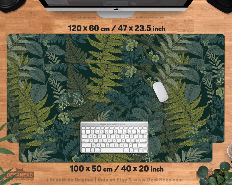 Pu&ograve; includere: Un grande tappetino da scrivania rettangolare con un design botanico con felci e altro fogliame in tonalit&agrave; di verde e blu. Il tappetino misura 120 x 60 cm e 100 x 50 cm. Una tastiera e un mouse bianchi sono sul tappetino.