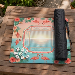 May include: A square mahjong mat featuring a tropical flamingo and hibiscus flower print in shades of pink, orange, and teal. The center includes game instructions and directional text. A black cylindrical carrying case sits on the right side.