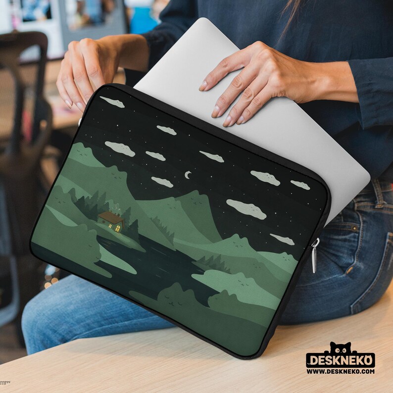 May include: A black laptop sleeve featuring a landscape design with mountains, trees, and a cabin under a starry night sky. The sleeve has a zipper closure and a black border. The laptop is being inserted into the sleeve.