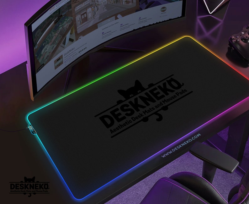 Op de afbeelding: Zwart rechthoekig DESKNEKO-muismat met een kleurrijke LED-rand. De mat heeft het DESKNEKO-logo en de tekst "Aesthetic Desk Mats and Mouse Pads". De website www.DESKNEKO.COM is ook zichtbaar.
