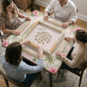 May include: A square mahjong game set featuring ivory-colored tiles with black, red, and green symbols, arranged on a rectangular play mat decorated with pink hibiscus flowers, green palm leaves, and small pagoda illustrations on a light background.