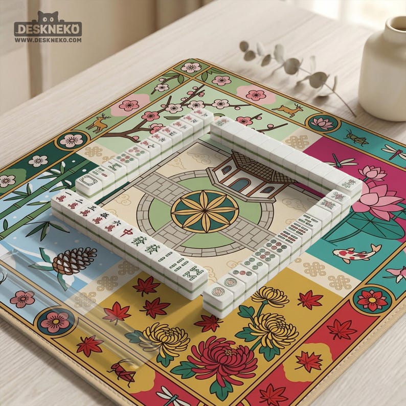 May include: A complete Mahjong set with white tiles and a colorful, floral-themed mat. The mat features various panels with illustrations of flowers, a pagoda, and a koi fish. The tiles are arranged in a square formation, ready for play.