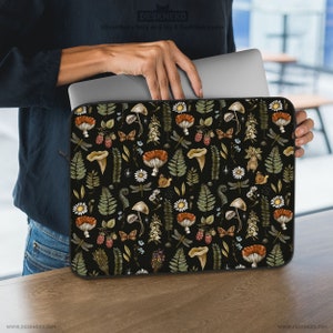 May include: A black laptop sleeve featuring a whimsical forest pattern. The design includes mushrooms, ferns, berries, butterflies, and dragonflies in shades of brown, green, and white. The sleeve is held by a person.
