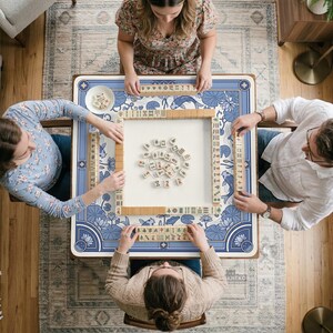 May include: A square mahjong table with a blue and white floral design is surrounded by four people. The table is set with tiles and a small dish. The game is in progress, with players holding tiles.