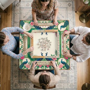 May include: A square mahjong table featuring a green border with peacock and pink flower illustrations. The center playing surface is cream-colored with scattered mahjong tiles, while four racks hold rows of tiles for each player at the table.