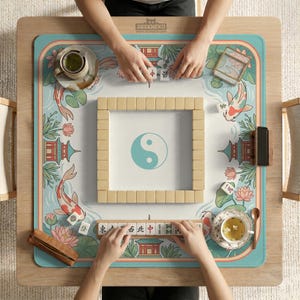 May include: A wooden mahjong table with a teal and white playing surface featuring koi fish, pagodas, and lotus flowers. The center has a yin-yang symbol. The game is in progress, with tiles and tea cups visible.