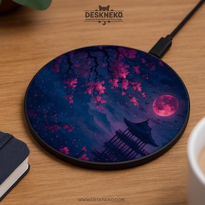 Puede incluir: Cargador inalámbrico redondo con un diseño de inspiración japonesa. El cargador presenta un fondo azul oscuro con una luna llena rosa, flores de cerezo y una pagoda. El cargador tiene el texto "Deskneko" y "www.deskneko.com".