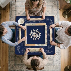 Puede incluir: Vista cenital de una partida de Mahjong en curso. Cuatro personas est&aacute;n sentadas alrededor de una mesa cuadrada azul marino con un dise&ntilde;o de peces koi y ruedas blancas. La mesa est&aacute; cubierta de fichas y rodeada de jugadores.