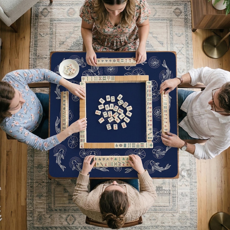 Puede incluir: Vista cenital de una partida de Mahjong en curso. Cuatro personas est&aacute;n sentadas alrededor de una mesa cuadrada azul marino con un dise&ntilde;o de peces koi y ruedas blancas. La mesa est&aacute; cubierta de fichas y rodeada de jugadores.