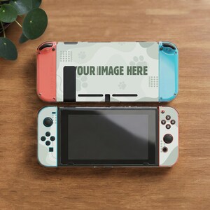 May include: A Nintendo Switch console with a custom skin featuring a light green and white camouflage pattern and the text "YOUR IMAGE HERE." The Joy-Con controllers are coral and teal. The console is on a wooden surface.