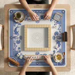 May include: Overhead view of a Mahjong game in progress. The game is set on a wooden table with a blue and white patterned mat featuring koi fish and lotus flowers. Beige tiles are arranged in a square, with players' hands visible. Tea cups and accessories are also present.