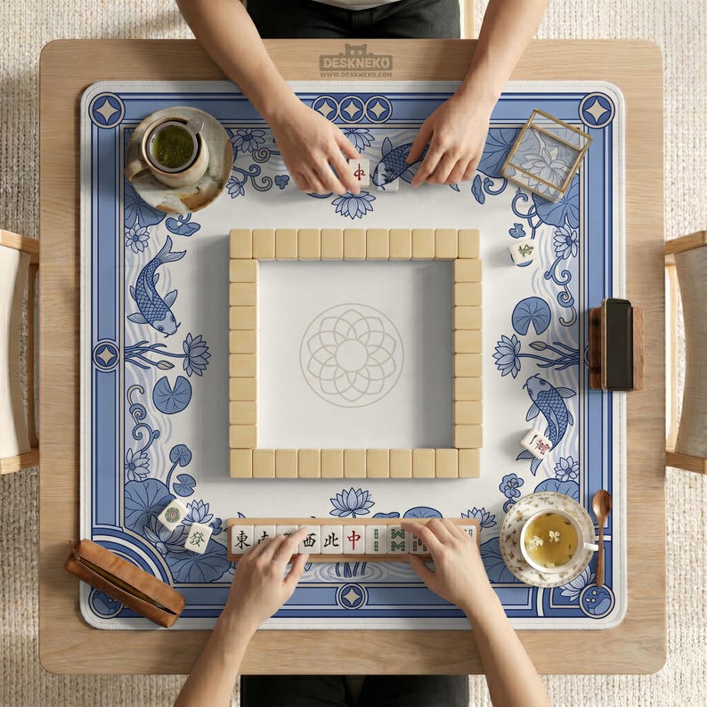 May include: Overhead view of a Mahjong game in progress. The game is set on a wooden table with a blue and white patterned mat featuring koi fish and lotus flowers. Beige tiles are arranged in a square, with players' hands visible. Tea cups and accessories are also present.