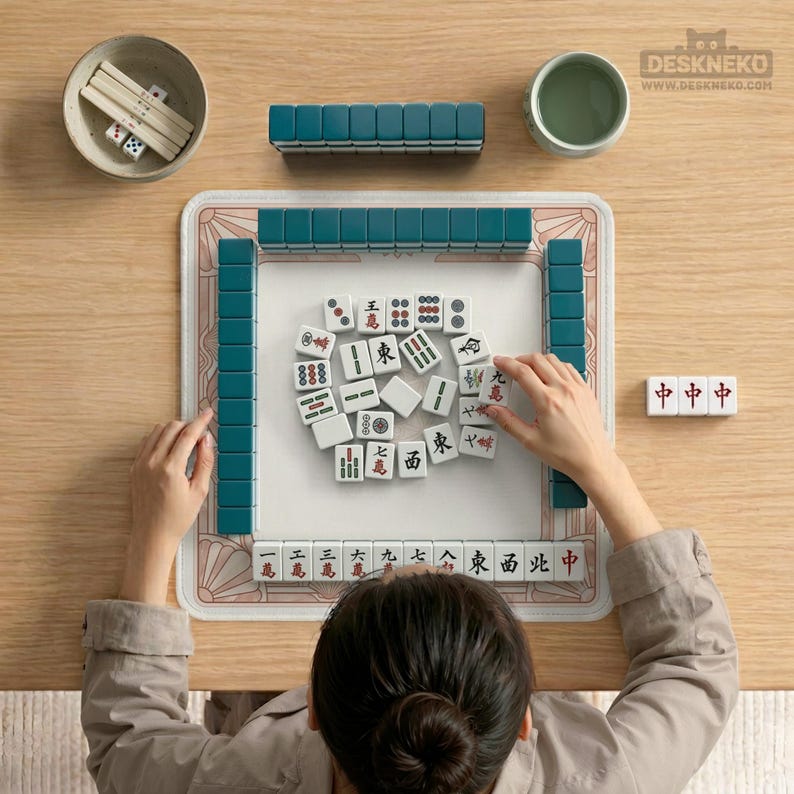 May include: A person playing Mahjong on a wooden table. The game set includes teal tiles, dice, and a bowl. The tiles are white with red and black symbols. A cup of tea is also on the table.