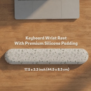 Pu&ograve; includere: Un poggiapolsi per tastiera con imbottitura in silicone, caratterizzato da una superficie grigio chiaro con un delicato motivo floreale botanico grigio scuro. Il prodotto misura 44,5 per 8,5 cm ed &egrave; posizionato sotto un portatile su una superficie in legno.