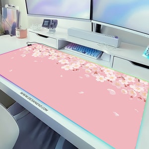 May include: A rectangular pink desk mat with a floral cherry blossom design and a glowing LED edge. The mat features the website address WWW.DESKNEKO.COM. The mat is on a white desk.