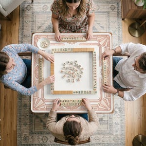 May include: Overhead view of a Mahjong game in progress. The game is set on a white table with a pink and beige art deco design. Four people are seated around the table, arranging tiles. The tiles are beige with black and red markings.