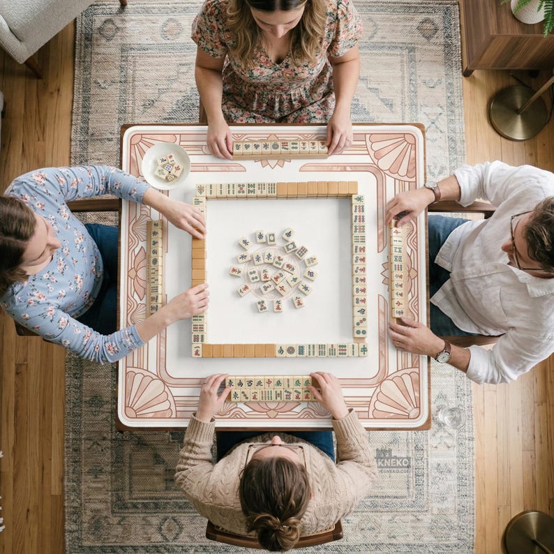 May include: Overhead view of a Mahjong game in progress. The game is set on a white table with a pink and beige art deco design. Four people are seated around the table, arranging tiles. The tiles are beige with black and red markings.