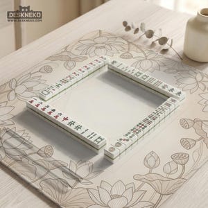 Puede incluir: Un juego de fichas de Mahjong blancas y verdes dispuestas en forma de cuadrado sobre una esterilla beige con dise&ntilde;os de flores de loto. Las fichas tienen caracteres y s&iacute;mbolos chinos. La esterilla est&aacute; sobre una superficie de madera clara.