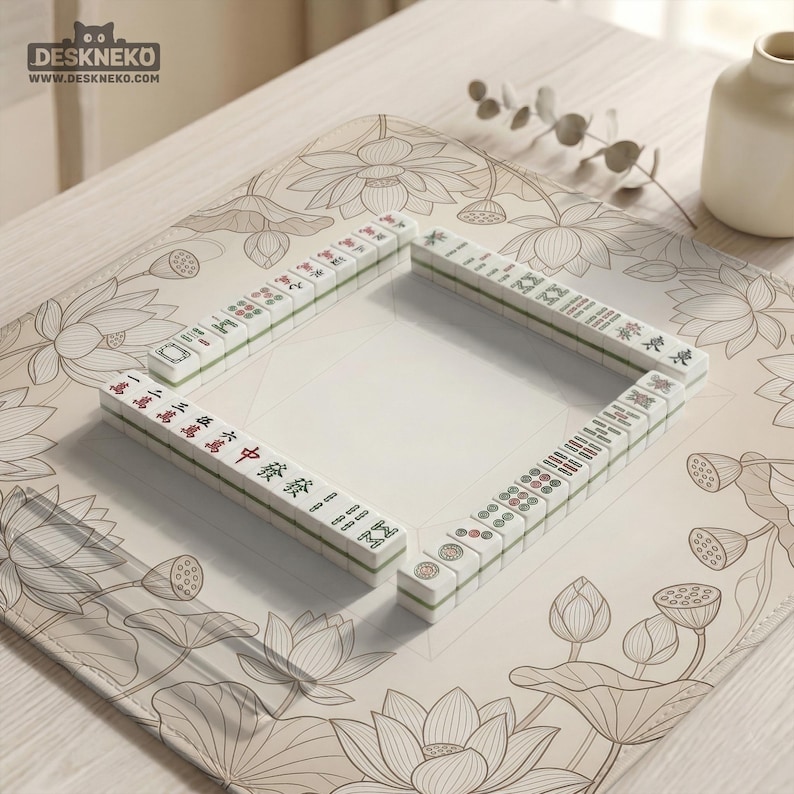 Puede incluir: Un juego de fichas de Mahjong blancas y verdes dispuestas en forma de cuadrado sobre una esterilla beige con dise&ntilde;os de flores de loto. Las fichas tienen caracteres y s&iacute;mbolos chinos. La esterilla est&aacute; sobre una superficie de madera clara.