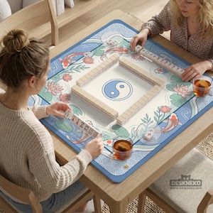 May include: A wooden square table with a Mahjong game mat. The mat has a blue border and a design of koi carp, lotus flowers, and a yin-yang symbol. Two people are playing Mahjong, with tiles and racks visible. Two cups of tea are on the table.