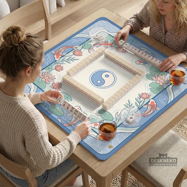 May include: A wooden square table with a Mahjong game mat. The mat has a blue border and a design of koi carp, lotus flowers, and a yin-yang symbol. Two people are playing Mahjong, with tiles and racks visible. Two cups of tea are on the table.