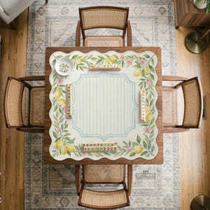 May include: Overhead view of a square wooden table with four cane-backed chairs. The table has a decorative mat with a lemon and floral design. The mat has a light blue and white striped center, and several game tiles are placed on the mat.