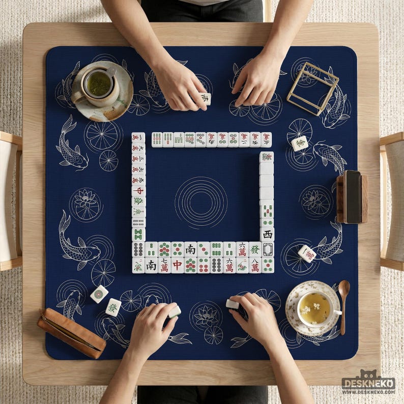 Puede incluir: Vista superior de un juego de Mahjong en curso sobre una mesa de madera. La alfombra azul marino presenta dise&ntilde;os de peces koi y flores de loto blancos. Se ven las manos de los jugadores, sosteniendo fichas. Tazas de t&eacute; y un soporte de fichas de madera.