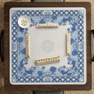 May include: A square mahjong game table with a blue and white floral design. The table features a white playing surface with a blue border, koi fish, and lotus flowers. Several mahjong tiles and a small bowl with dice are visible.