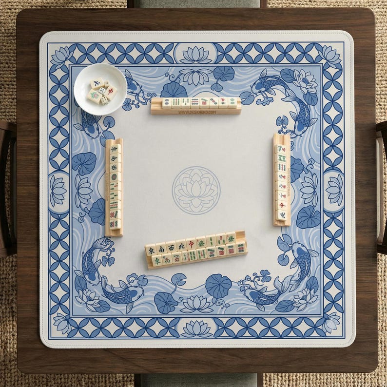 May include: A square mahjong game table with a blue and white floral design. The table features a white playing surface with a blue border, koi fish, and lotus flowers. Several mahjong tiles and a small bowl with dice are visible.