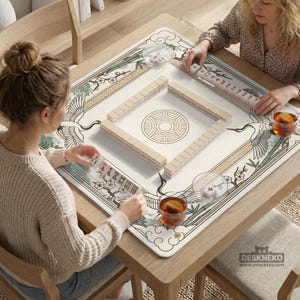 May include: A square mahjong game table with a cream-colored playing surface featuring a crane and floral design. The table is made of wood and has a decorative border. Two people are playing mahjong, with tiles and tea cups visible.