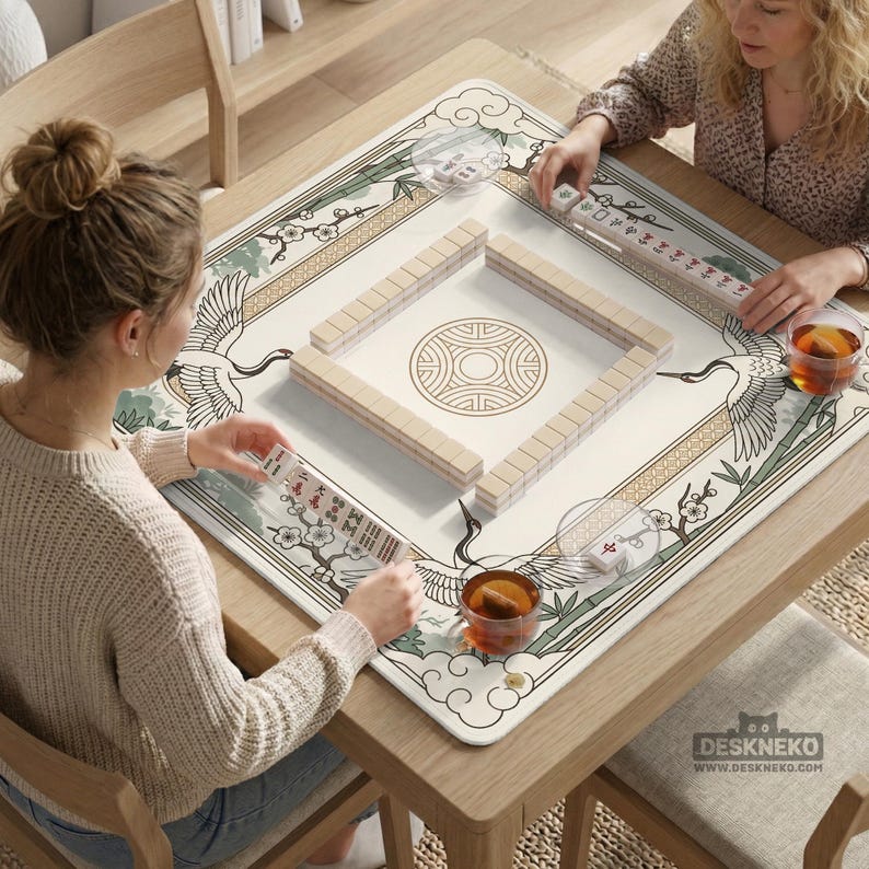 May include: A square mahjong game table with a cream-colored playing surface featuring a crane and floral design. The table is made of wood and has a decorative border. Two people are playing mahjong, with tiles and tea cups visible.
