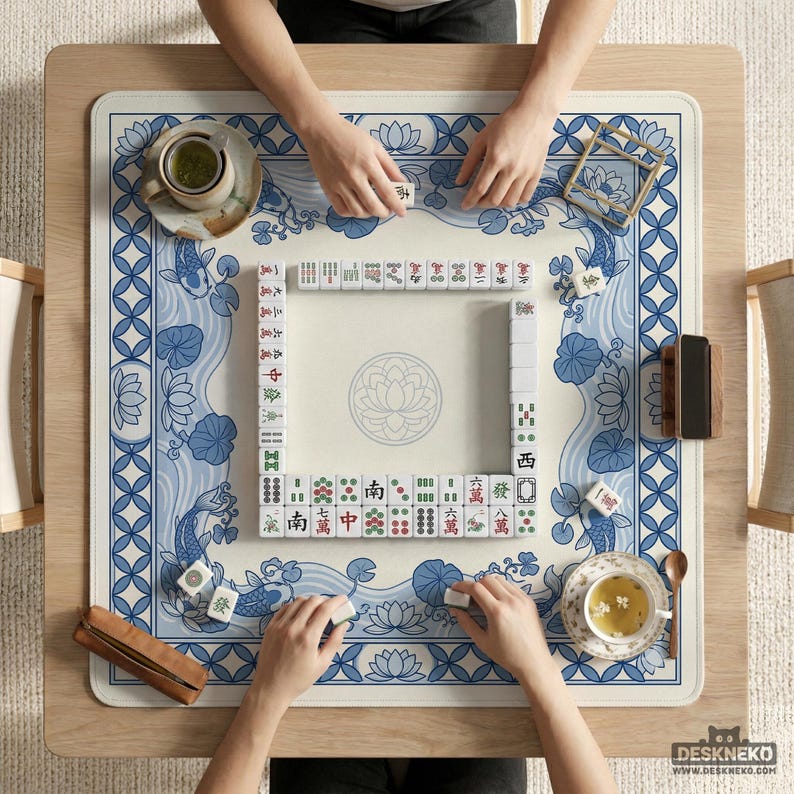 May include: Overhead view of a Mahjong game in progress. The game is set on a light wood table with a blue and white patterned mat. The mat features koi fish and lotus flower designs. Players are arranging tiles, with tea cups and accessories nearby.