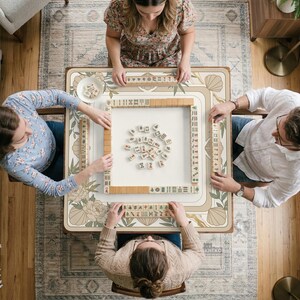 May include: Overhead view of a Mahjong game in progress. Four people are seated around a square table with a floral design. The table is set with tiles and racks. The game is in play, with tiles scattered in the center.