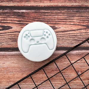 Peut inclure: Un emporte-pièce en silicone bleu clair en forme de manette de jeu vidéo. L'emporte-pièce est posé sur une surface en bois avec une grille en fil métallique noire.