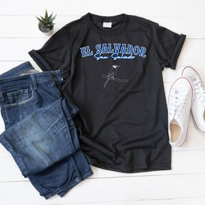 Peut inclure: T-shirt noir avec l'inscription "EL SALVADOR San Salvador" en bleu et blanc. Un dessin au trait blanc d'un oiseau perché sur une branche se trouve en dessous du texte. Le t-shirt est associé à un jean bleu et des baskets blanches.
