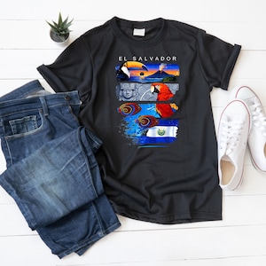 Peut inclure: T-shirt noir avec un motif graphique coloré représentant le texte "EL SALVADOR" et des images d'un toucan, d'un volcan, d'une statue maya, d'un perroquet et du drapeau du Salvador. Le t-shirt est associé à un jean bleu et à des baskets blanches.