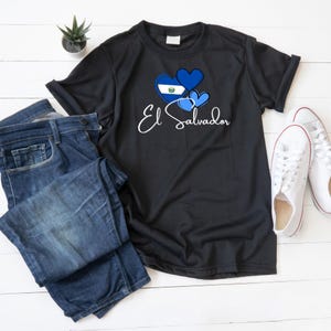 May include: Black t-shirt with a blue heart design featuring the flag of El Salvador and the text "El Salvador".