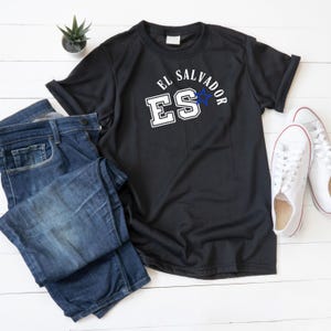 Peut inclure: T-shirt noir avec l'inscription "EL SALVADOR" et "ES" en lettres blanches avec une étoile bleue. Le t-shirt est associé à un jean bleu et des baskets blanches sur une surface en bois blanc. Une petite plante verte est également présente.